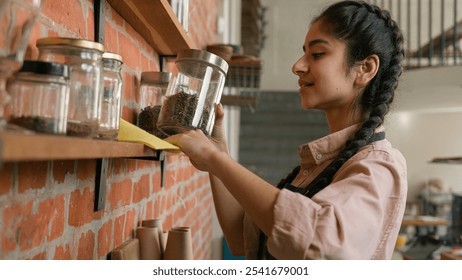 Indian woman waitress barista in apron wipe dust using cleaning duster arabian smiling female girl cleaner bar employee wiping washing shelf with spices in cafe restaurant clean shelves in cafeteria - Powered by Shutterstock