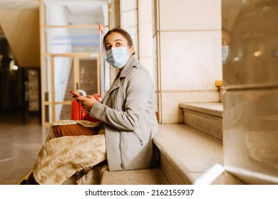 Indian Woman Wearing Face Mask Using Cellphone While Sitting On Stairs In Airport Indoors