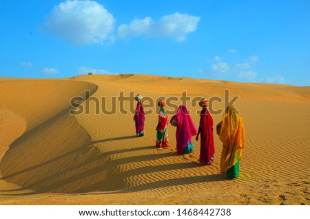 Indian women carrying heavy jugs of water on their head and walking on a yellow sand dune in the hot summer desert against blue sky.water crises, jaisalmer, rajasthan, india