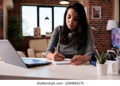 Indian Worker Taking Paperwork Notes On Notebook, Writing Information Report Or School Class Lesson. Working On Laptop With Textbook To Do Research Online And Plan Web Presentation.