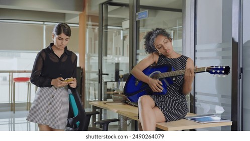 Indian young adult gen z bored business woman use mobile phone do online chat typing message standing indoor job office. Teen curly hair girl sitting on desk table trying to play guitar at work place - Powered by Shutterstock