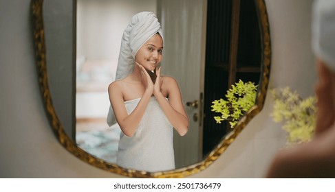 Indian young adult gen z beautiful woman wrapped towel on head and body after shower reflected look bathroom mirror use cosmetics beauty product apply moisturizer cream on face do skin care routine - Powered by Shutterstock