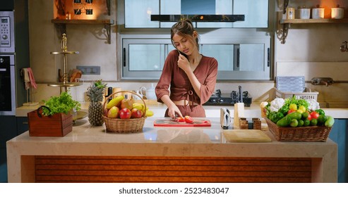 Indian young adult gen z beautiful woman standing use mobile answer phone call talking casual gossip distant conference with friend hold knife cut tomato slice on chopping board indoor modern kitchen - Powered by Shutterstock