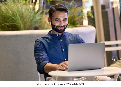 Indian Young Business Man Wearing Earbud Having Virtual Meeting Online Remote Conference Video Call Looking At Laptop Computer Remote Working Watching Webinar Sitting In Night City Cafe Or Park.