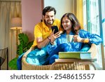 Indian young couple happily chatting on a video call while seated by a sunlit window, sharing joyful moments together with a smartphone in cozy setting