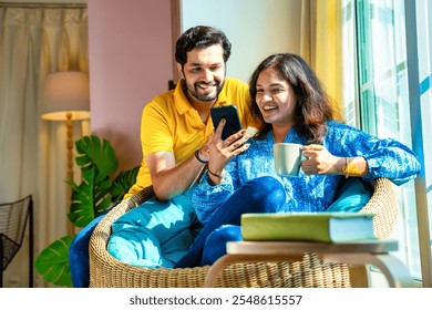 Indian young couple happily chatting on a video call while seated by a sunlit window, sharing joyful moments together with a smartphone in cozy setting - Powered by Shutterstock