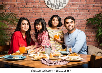 Indian Young Family Of Four Eating Food At Dining Table At Home Or In Restaurant. South Asian Mother, Father And Two Daughters Having Meal Together