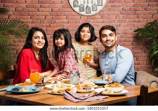 Indian young Family of four eating food at\
dining table at home or in restaurant. South Asian mother, father\
and two daughters having meal\
together
