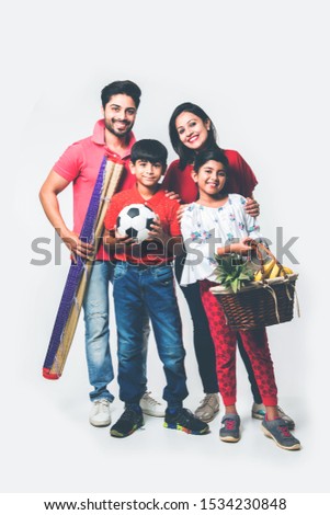 Indian young Family of four going for Picnic - standing over white background with fruit basket, mat, football and drinks. selective focus