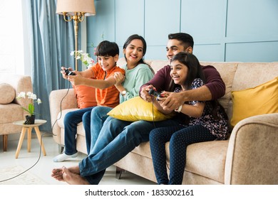 Indian Young Family Of Four Playing Video Game Using Controller Or Joystick While Sitting On Sofa