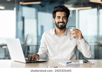 An Indian young male businessman is sitting in the office at his desk and holding a glass of clean water in his hand. He looks at the camera with a smile. - Powered by Shutterstock