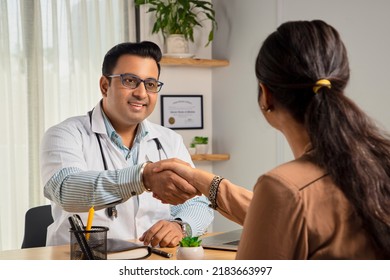 An Indian Young Man Or Male Smiling Physician Or Doctor Sitting In A Modern Clinic Wearing A Stethoscope And Apron Shaking Hand With A Happy Female Patient. Medical, Medicine, And Healthcare Concept