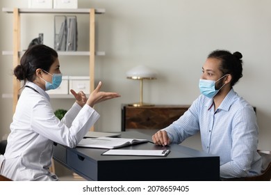 Indian Young Woman Doctor Physician In Protective Medical Mask Consulting African American Man Patient At Meeting In Hospital, Discussing Checkup Results Or Symptoms, Coronavirus Concept