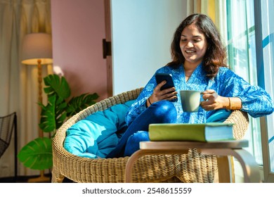 Indian young woman enjoying video call on smartphone with coffee mug in hand, sitting by sunlit window in cozy chair, smiling in warm morning light - Powered by Shutterstock