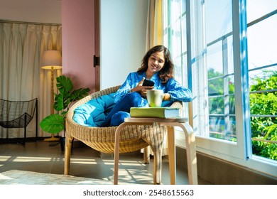 Indian young woman enjoying video call on smartphone with coffee mug in hand, sitting by sunlit window in cozy chair, smiling in warm morning light - Powered by Shutterstock