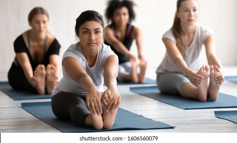 Indian young woman practicing yoga at group lesson, diverse people doing Seated forward bend exercise on mats, stretching in paschimottanasana pose, working out in modern yoga center - Powered by Shutterstock