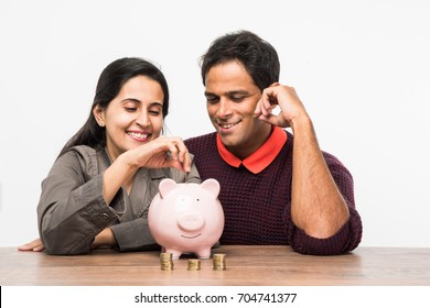 Indian/Asian Smart And Young Couple Saving Money In Piggy Bank For Future While Sitting Across Table