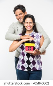 Indian/Asian Young Couple Buying / Dreaming For Home OR Real Estate Concept, Holding 3D Colourful Paper Model Of House. Isolated Over White Background
