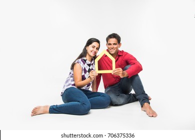 Indian/Asian Young Couple Buying / Dreaming For Home OR Real Estate Concept, Holding 3D Colourful Paper Model Of House. Isolated Over White Background
