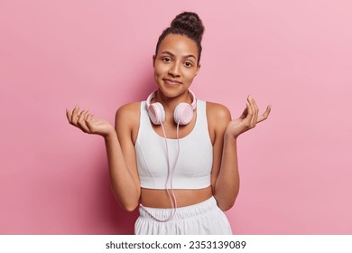 Indifferent hesitant sporty woman spreads palms and shrugs shoulders doesnt know exact time when training starts expresses uncertainty wears white top shorts uses headphones poses over pink background - Powered by Shutterstock