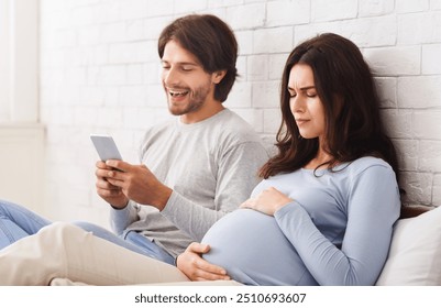 Indifferent husband. Future father using smartphone ignoring his pregnant wife that suffering belly ache on bed at home, free space