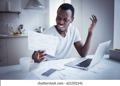 Indignant Angry Irritated Young African American Man Looking At Utility Bill, Frustrated About High Taxes, Worried About Troubles With Mortgage Payment To Bank, Having Serious Problems With Debt