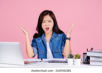 Indignant  Asian business woman wear casual shirt sit work at white office desk. She scream and spread hands isolated on pink background
