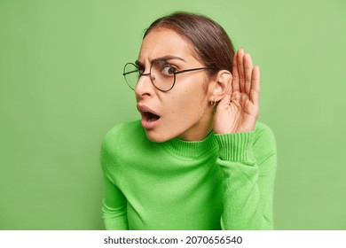 Indignant brunette woma tries to overhear someone holds hand near ear tries to understand words eavesdropping keeps mouth opened wears spectacles and poloneck isolated over green background.