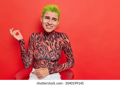 Indignant displeased young woman with green trendy hairstyle raises hand looks unhappily frowns face wears turtleneck poses against pink background. Fashionable hipster girl has unique appearance.