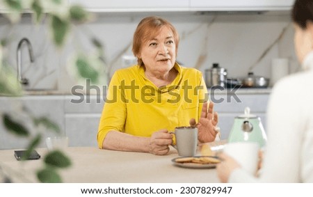 Indignant elderly woman with cup of tea sits in kitchen and listens to daughter.Women talk about family difficulties.Concept of troubled family relationships