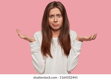 Indignant lovely woman shruggs shoulders, has hesitant expression, dressed in casual white sweater, expresses apathy, isolated over pink background, feels clueless about her future. Puzzlement concept