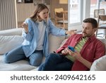 Indignant, offended, hysterical woman screams at husband sitting on sofa with cellphone. Couple family quarrel conflict misunderstanding discord. Indifferent man ignores dissatisfied wife