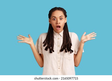 Indignant shocked woman with black dreadlocks has surprised angry expression, raised arms, looking at camera with big eyes, wearing white shirt. Indoor studio shot isolated on blue background.
