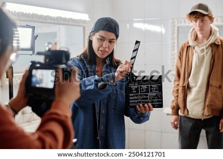 Individual holding a clapboard, directing scene on film set, with observer watching preparation closely. Camera operator ready for filming in indoor setup