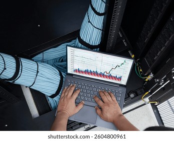 Individual performing data analysis on a laptop in a server room, surrounded by organized cables. The screen displays performance graphs and statistics, highlighting important trends.