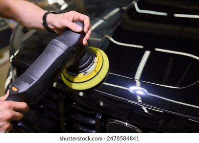 An individual is using a machine to polish the black cars hood, bumper, rims, automotive mirror, and other exterior automotive parts