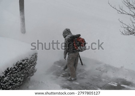 Individual using a powerful backpack leaf blower to clear fresh snow from a paved residential sidewalk during a heavy winter snowfall, showcasing arduous winter maintenance.