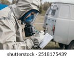 An individual wearing protective gear and a gas mask writes notes on a pad while standing next to a white vehicle in a potentially hazardous area.