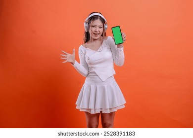 indonesia woman in white sweater happy wearing earphone headphone, holding mobile phone screen, cellphone green screen while waving sideways. for entertainment, lifestyle, business, service concept - Powered by Shutterstock