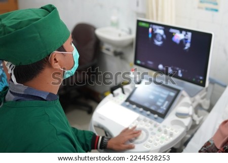 Indonesian doktor observes the patient with a fetal detection device. An obstetrician wearing  sterile one-off suit and scrub cap watching the screen of ultrasonographic diagnosis.