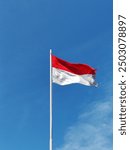 The Indonesian flag flutters on a pole blown by the wind against a background of blue sky and clouds.