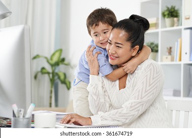 Indonesian Little Boy Hugging His Mom, Who Is Computing In Home Office