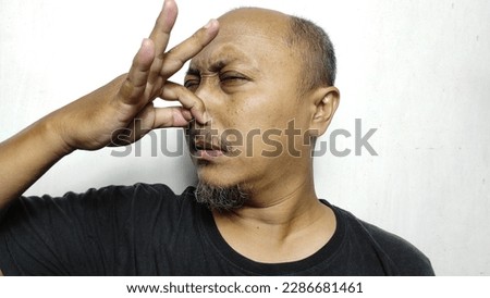 An Indonesian man with a bald head, wearing a black t-shirt against a white background, was pinching his nose because he couldn't stand the pungent smell