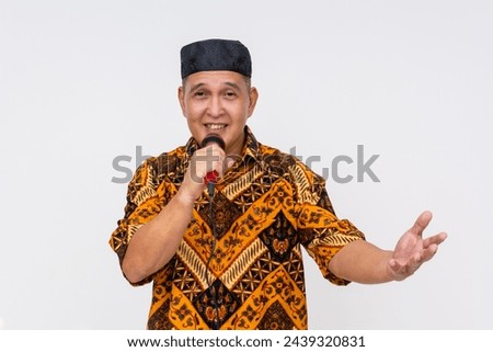 An Indonesian man in a traditional batik shirt and kopiah hat emceeing, with a microphone, isolated on white.