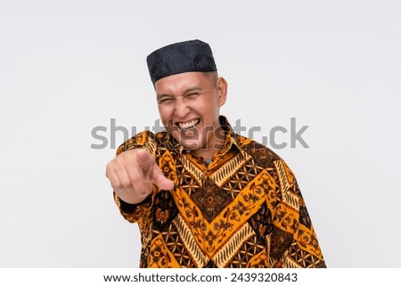 Indonesian man wearing a batik shirt and a kopiah hat chuckling poking fun at someone. Isolated on a white background.