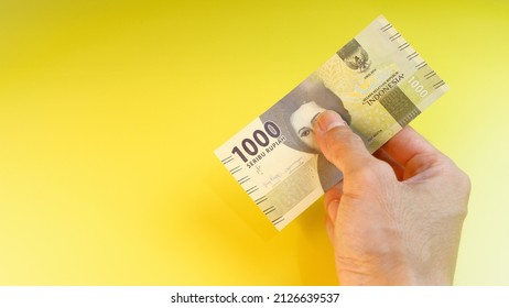 Indonesian Rupiah the official currency of Indonesia. A man's hand is making a payment. Male hand showing Indonesian Rupiah note. Business and finance concept. Yellow Background. Uang 1000 Rupiah. - Shutterstock ID 2126639537
