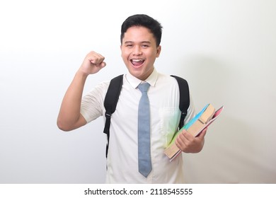 Indonesian Senior High School Student Wearing White Shirt Uniform With Gray Tie Holding Some Books, Smiling And Raising His Fist, Celebrating Success. Isolated Image On White Background