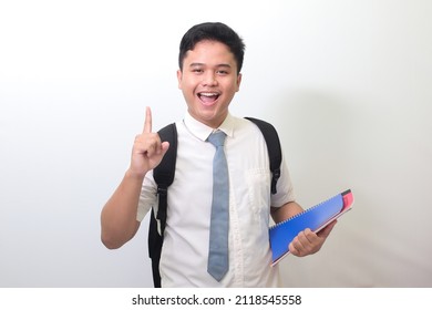 Indonesian Senior High School Student Wearing White Shirt Uniform With Gray Tie Holding Some Books, Smiling And Pointing Up With Finger Reminding Something. Isolated Image On White Background