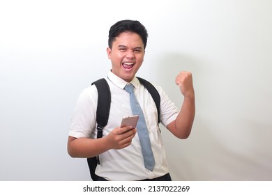 Indonesian Senior High School Student Wearing White Shirt Uniform With Gray Tie Raising His Fist, Celebrating Success While Holding A Mobile Phone. Isolated Image On White Background