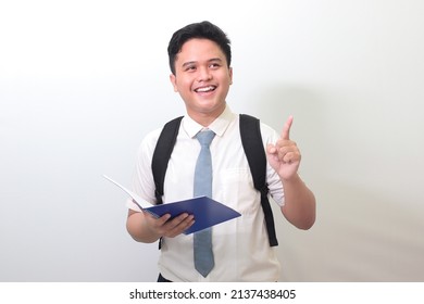 Indonesian Senior High School Student Wearing White Shirt Uniform With Gray Tie Pointing At Empty Space While Holding An Opened Note Book. Isolated Image On White Background